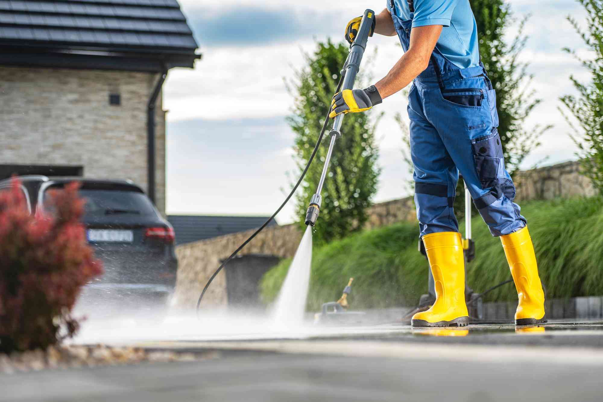Worker in yellow boots pressure washing a driveway in front of a house