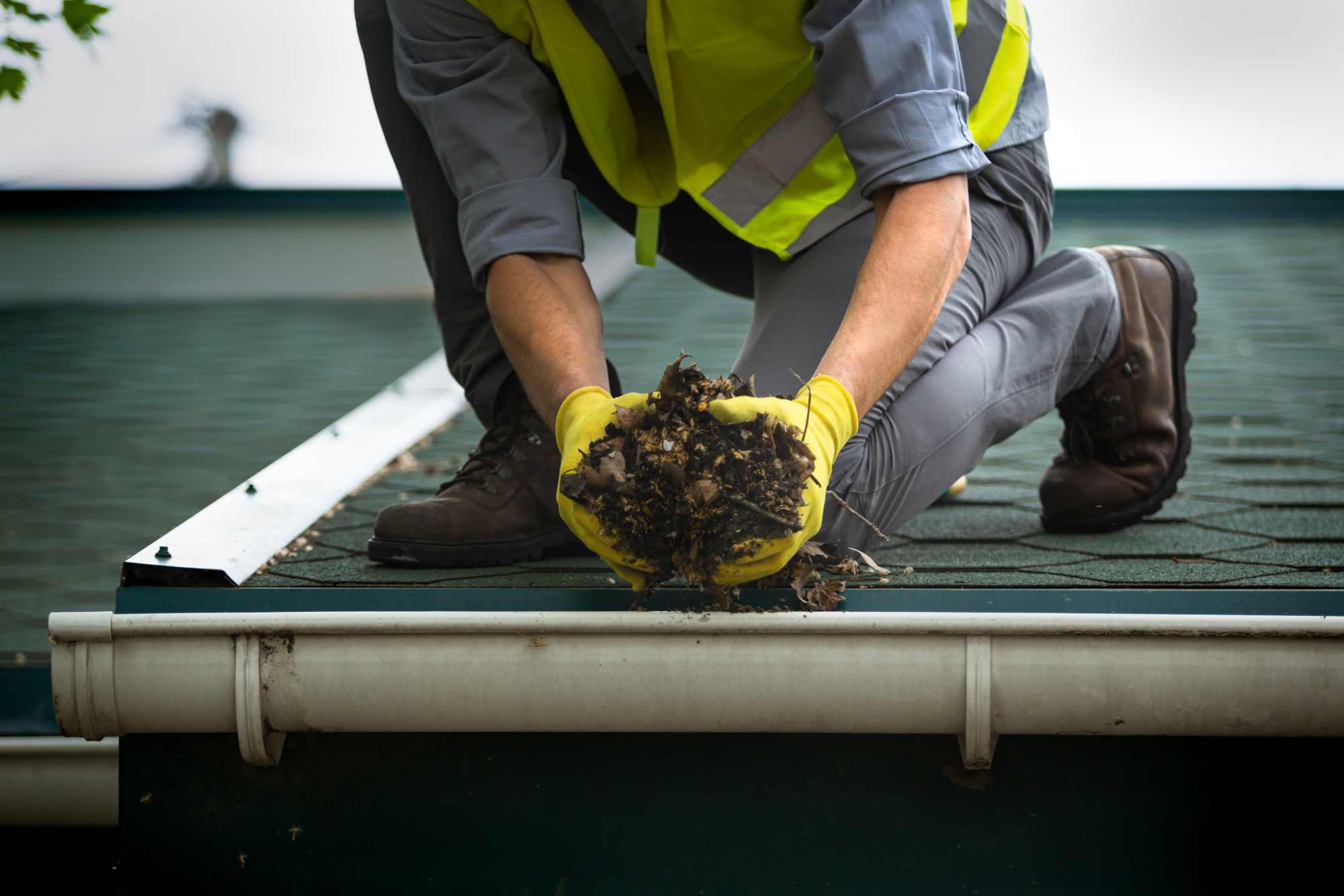 Worker in a hi-vis vest lifting leaves and debris out of a roof gutter