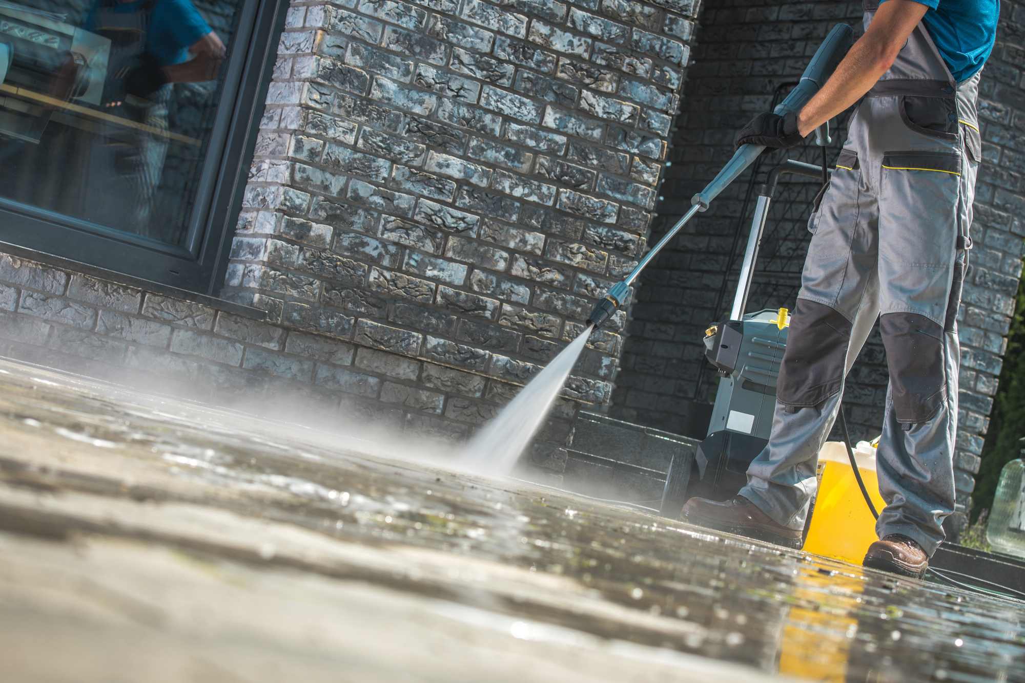 Worker pressure cleaning paving next to a brick wall